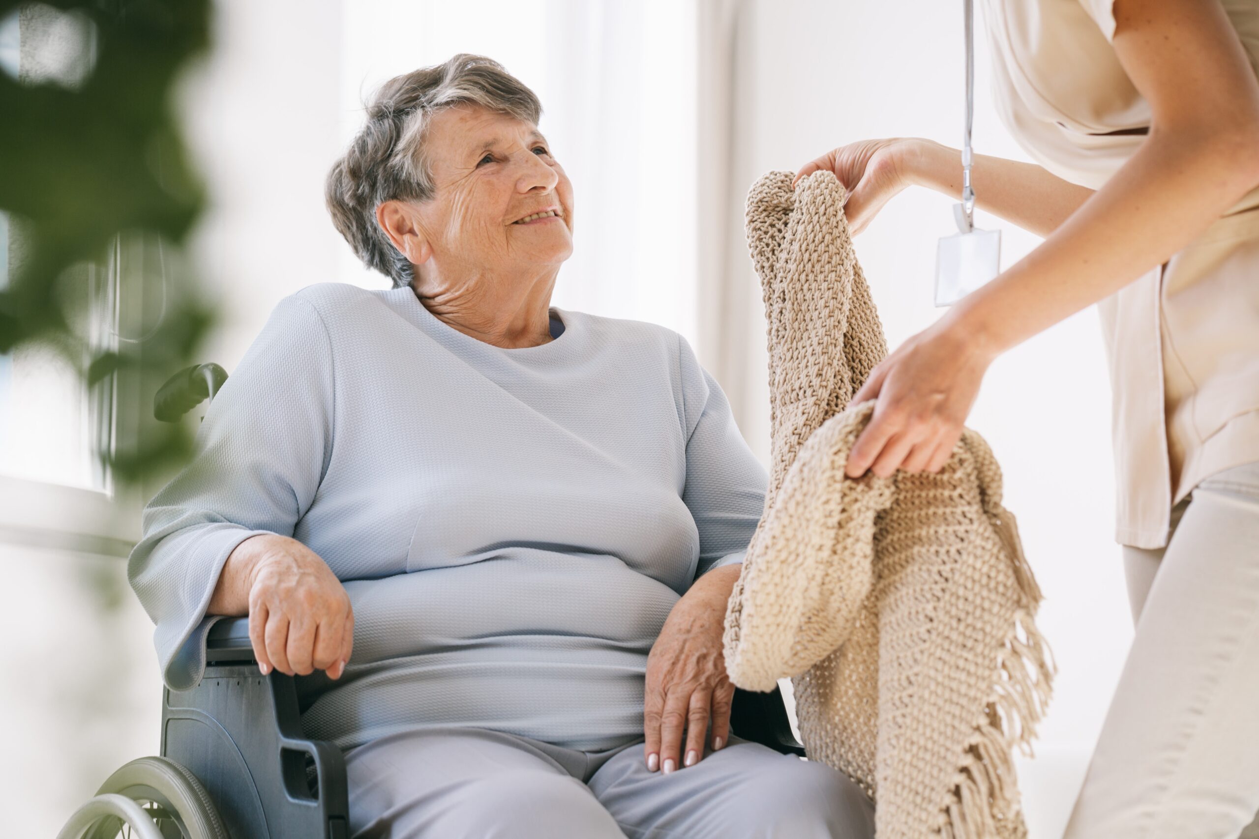 Ältere Dame im Rollstuhl lächelt eine Pflegekraft an, die ihr eine Strickdecke in Zolling oder Freising reicht