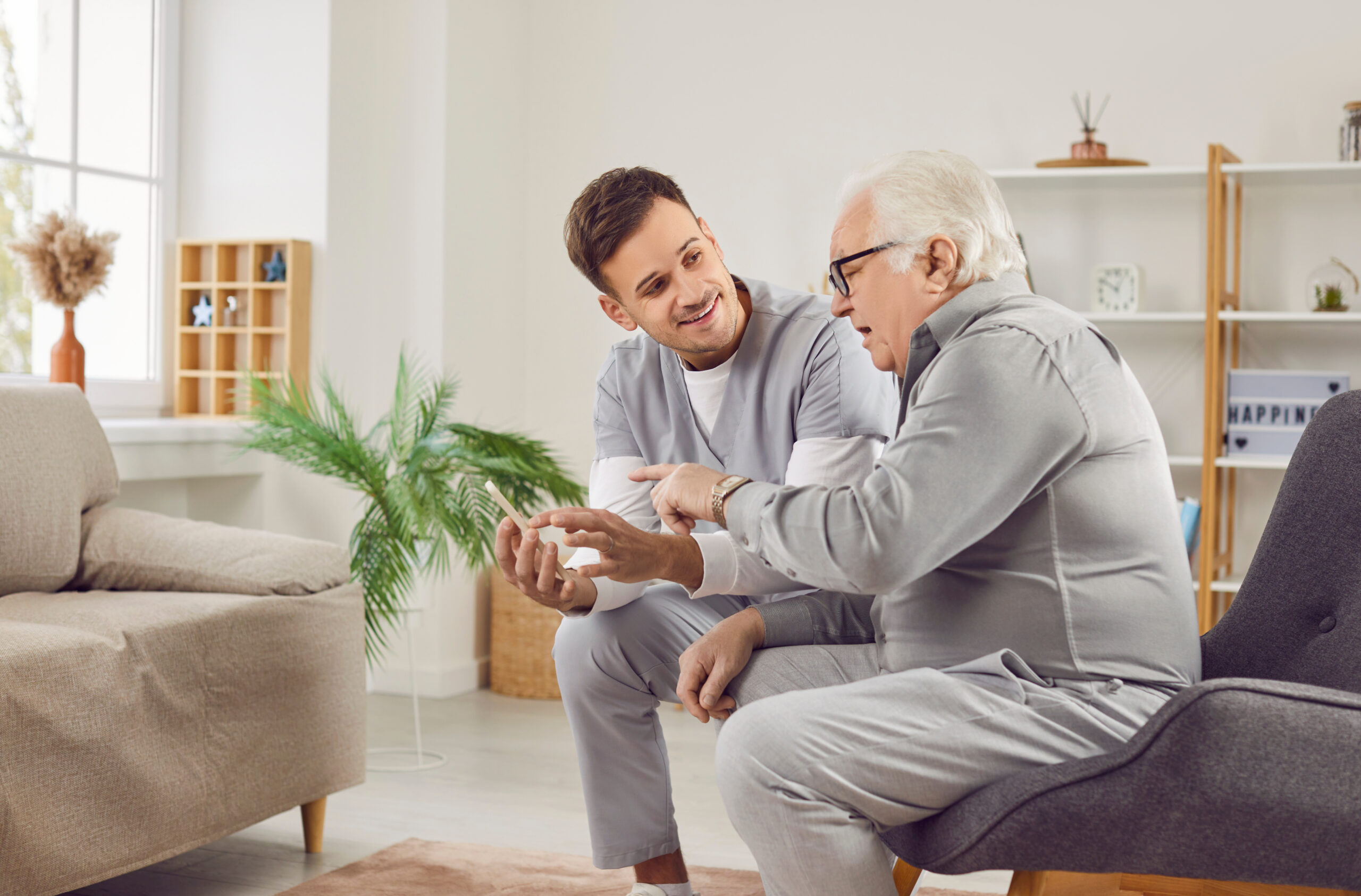 Pflegekraft sitzt mit älterem Mann im Wohnzimmer und erklärt ihm Familienpflegezeit auf einem Smartphone