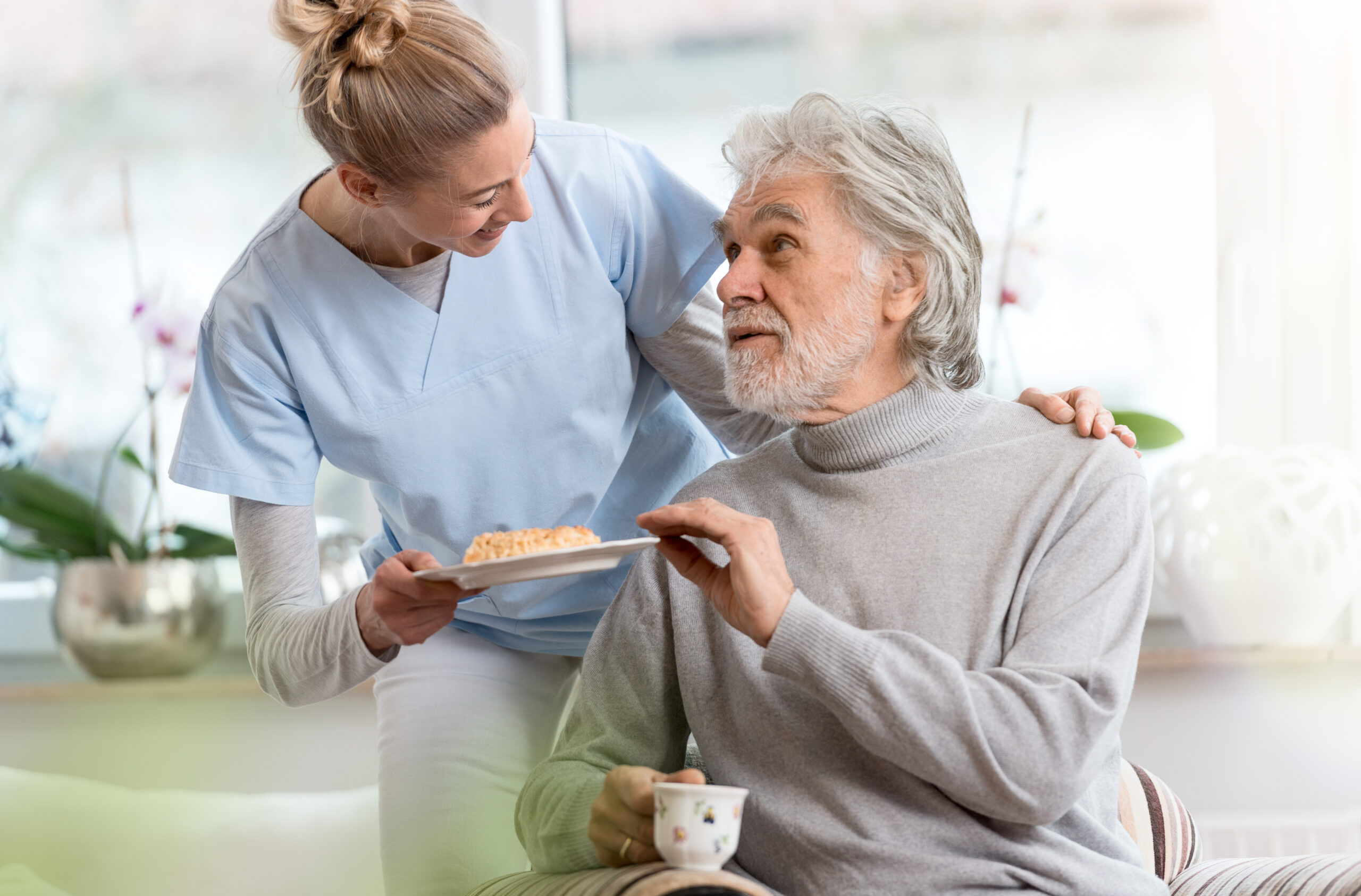 Pflegekraft bringt einem älteren Mann Kuchen und lächelt ihn selbstbestimmt freundlich an